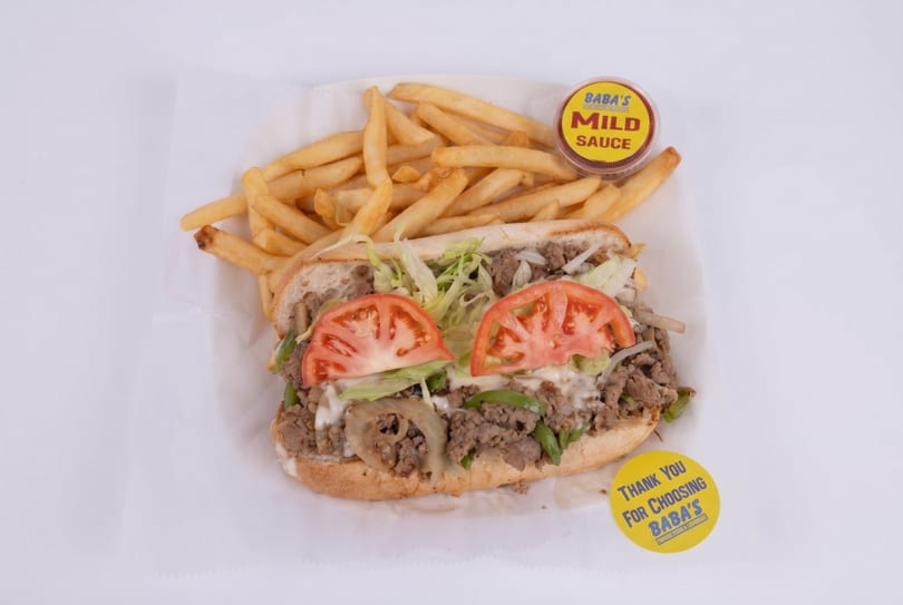 A Baba's cheesesteak with lettuce, tomatoes, onions, and peppers, served with fries and mild sauce.